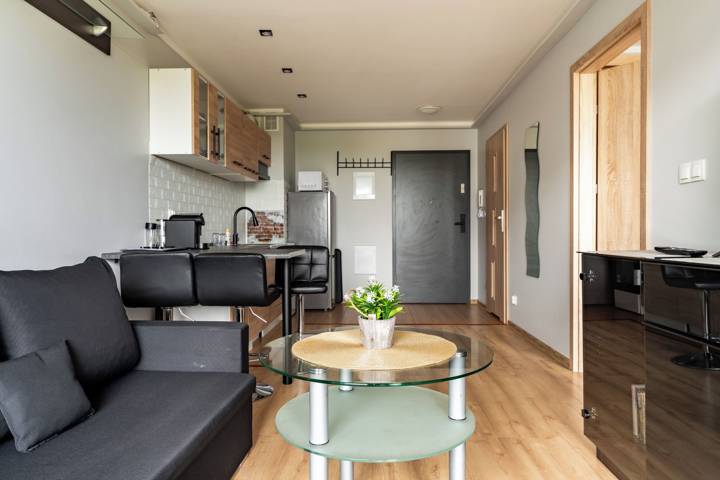 A modern living room with a glass coffee table, a black sofa, and a kitchen in the background.