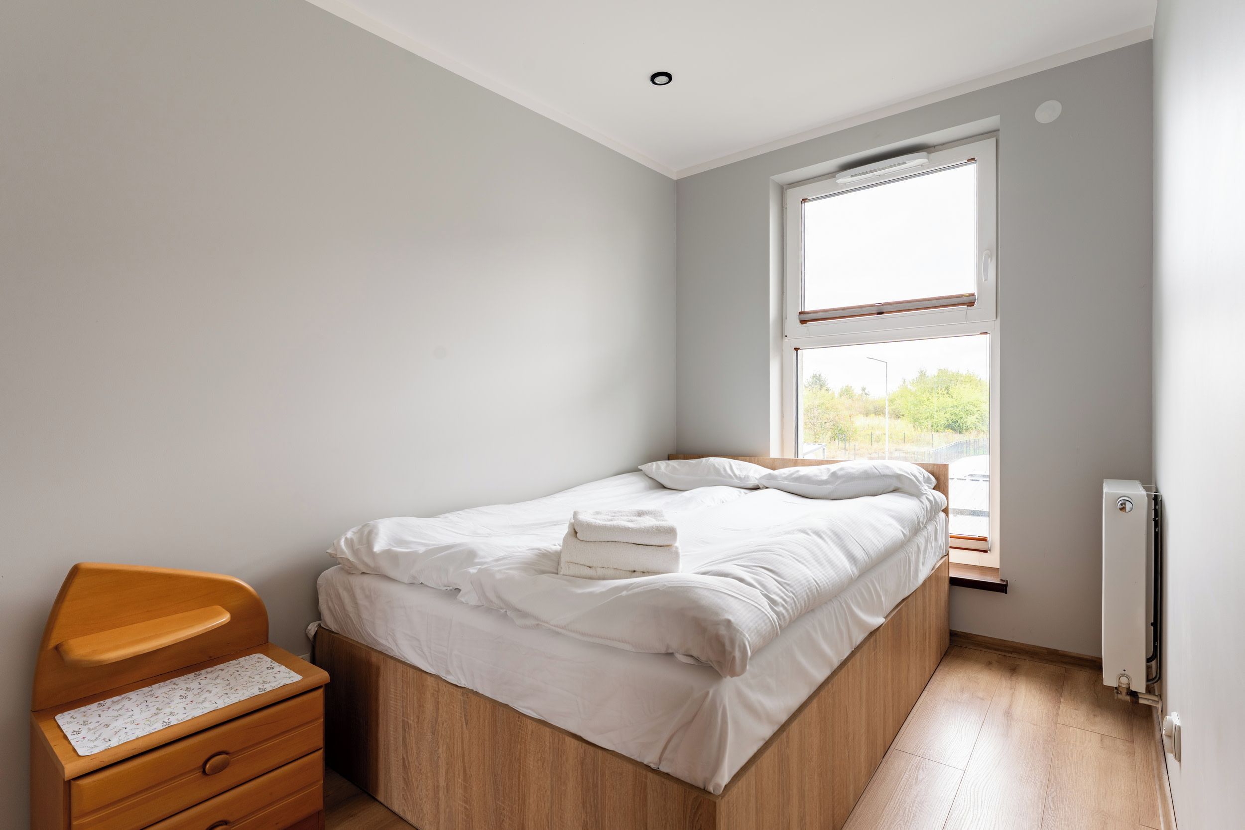 A bedroom with a single bed, a wooden nightstand, a window with blinds, and a radiator.
