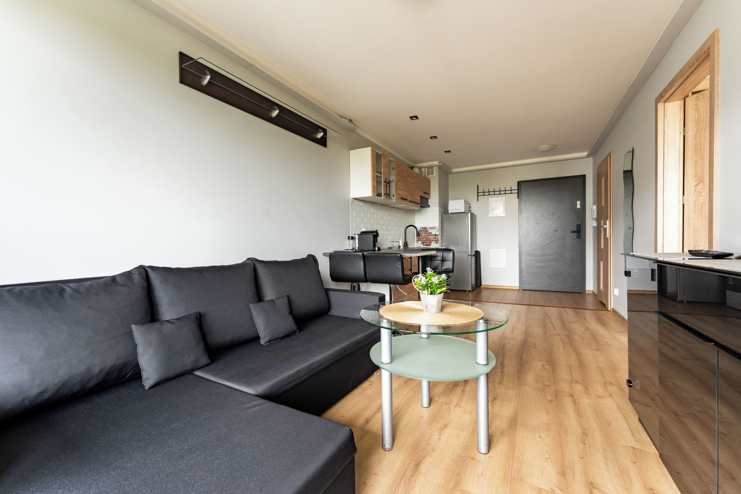 Una sala de estar moderna con un sofá negro, una mesa de centro de vidrio y una cocina al fondo.