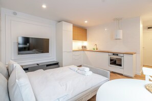 Ein modernes Hotelzimmer mit Flachbildfernseher, einem Bett mit weißer Bettwäsche, einer Küchenzeile mit Mikrowelle, Backofen und Spüle sowie einer kleinen Sitzecke.