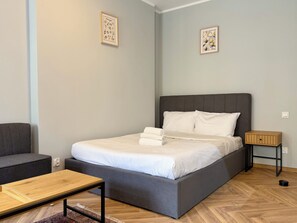 Ein modernes Schlafzimmer mit einem grau gepolsterten Bett, weißen Bettwäsche, einem hölzernen Nachttisch und einem grauen Sofa.