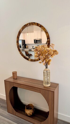 A wooden console table with a vase of dried leaves and a candle, a round mirror on the wall, and a kitchen in the background. - Modern Cozy Retreat Minutes From Downtown Houston (Houston)