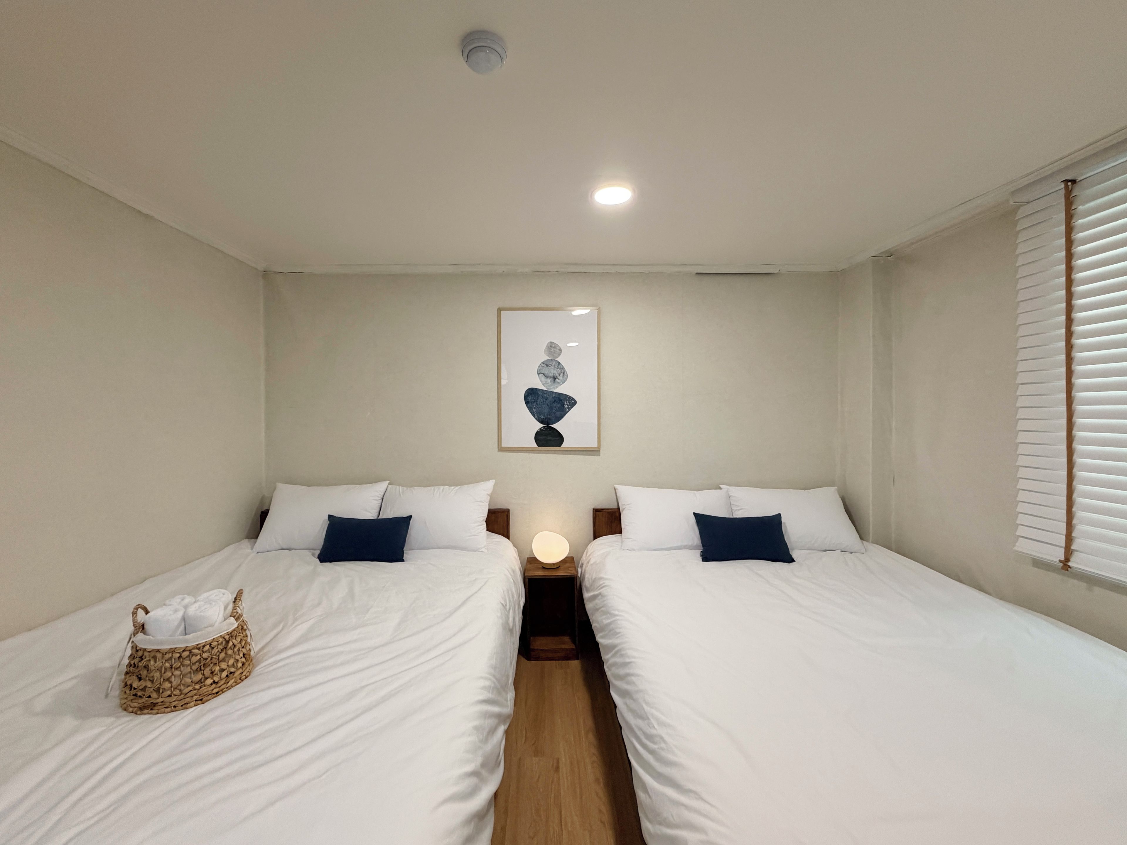Habitación de hotel con dos camas, una mesita de noche con lámpara, una obra de arte en la pared y una ventana con persianas.