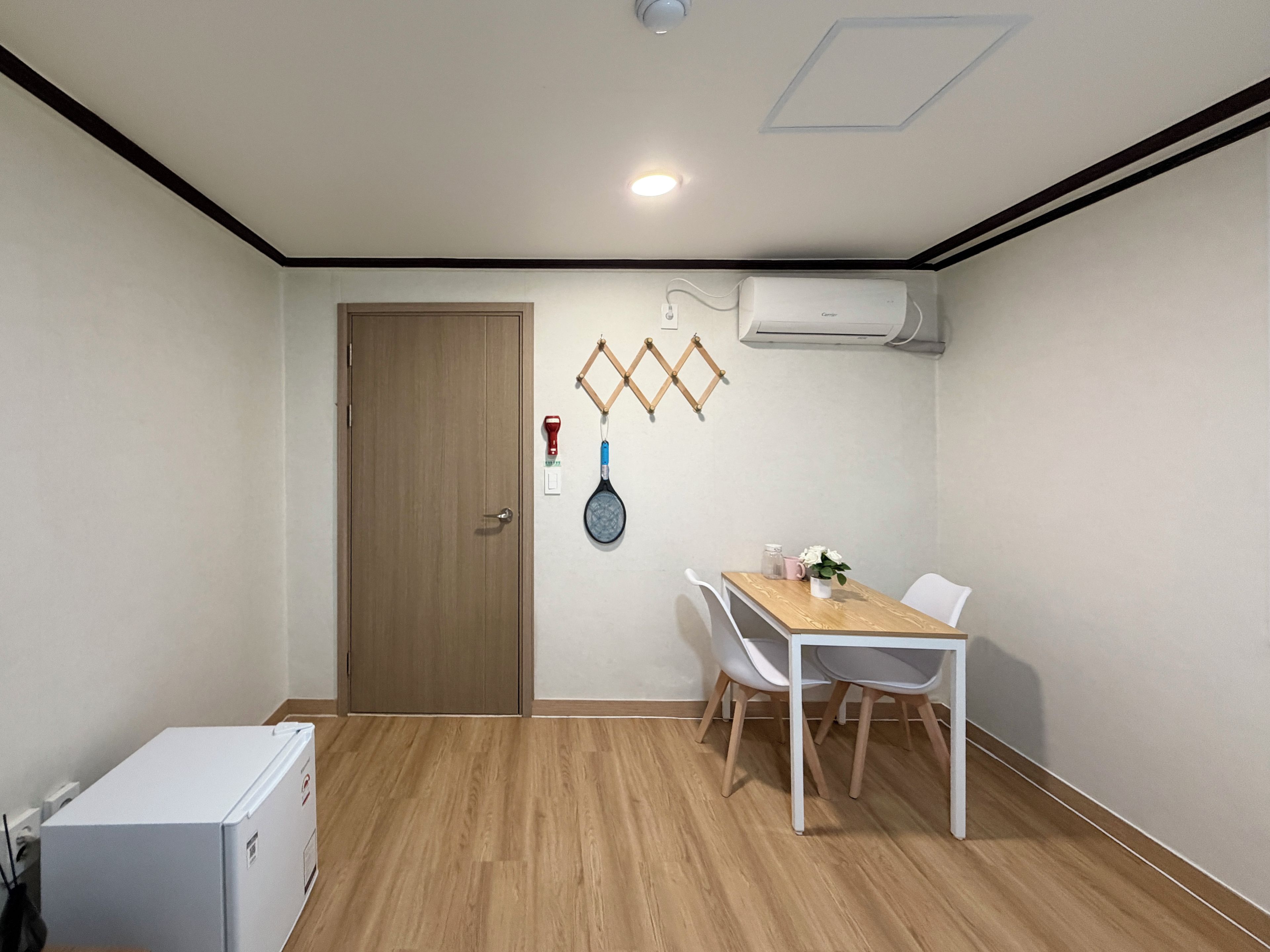 Una habitación con suelo de madera, una mesa blanca con sillas, un refrigerador blanco y un equipo de aire acondicionado en la pared.