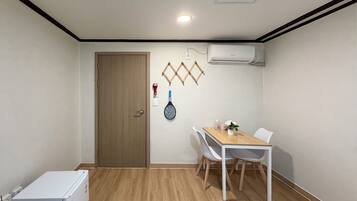 Una habitación con suelo de madera, una mesa blanca con sillas, un refrigerador blanco y un equipo de aire acondicionado en la pared.