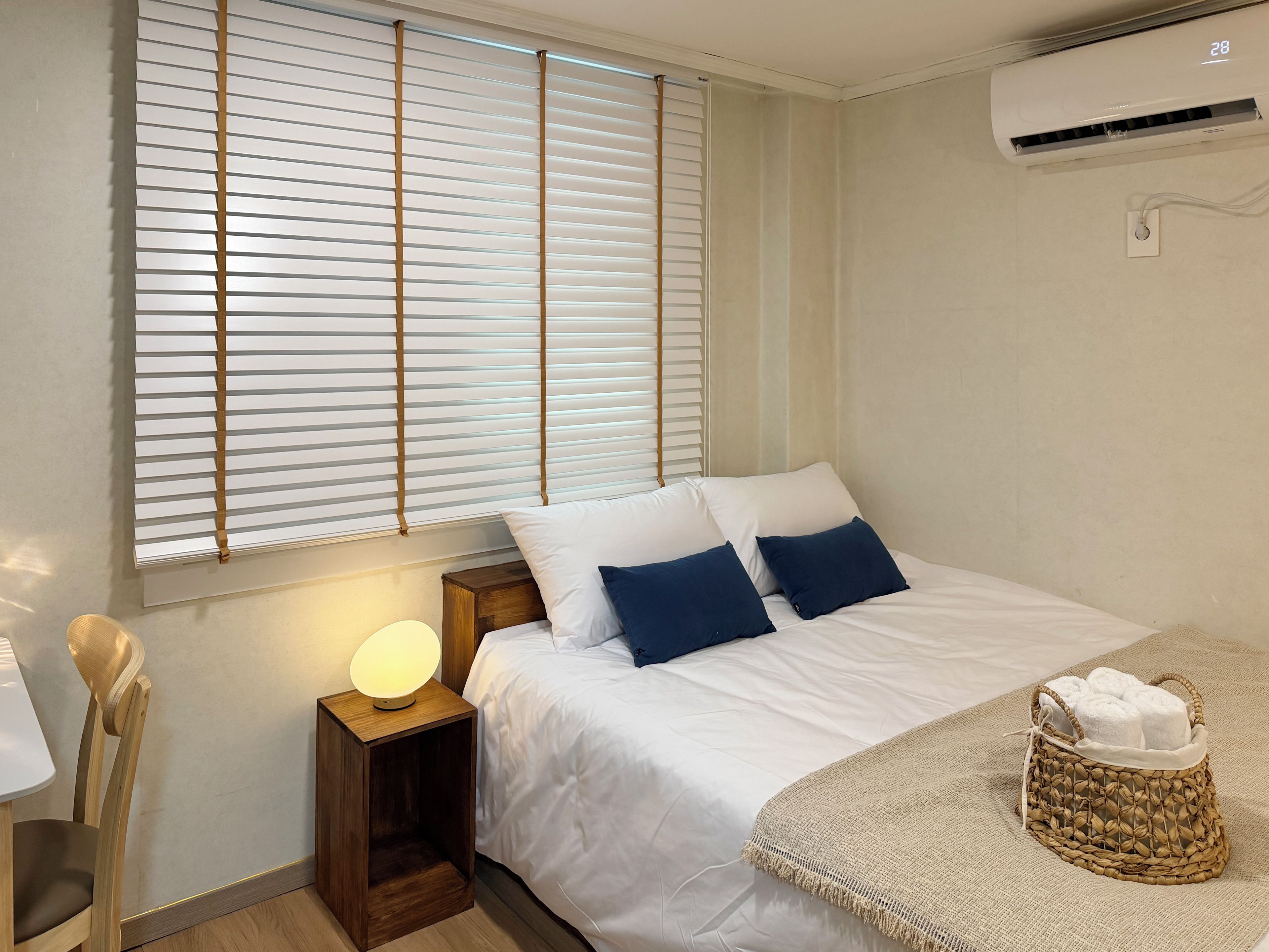 A bedroom with a bed, bedside table, lamp, and a window with blinds.