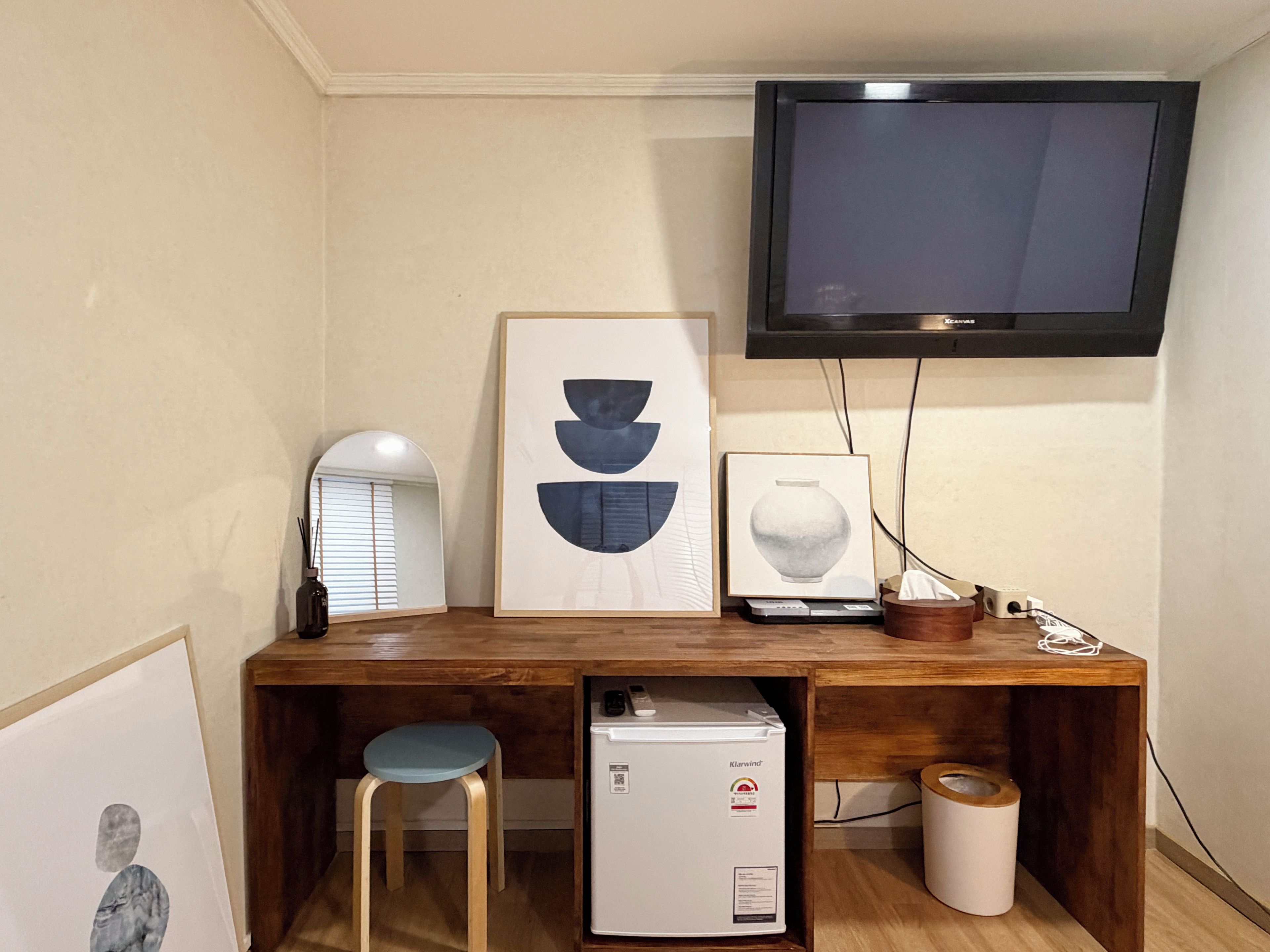 A compact room with a wooden desk, a small white refrigerator, a television mounted on the wall, and a framed abstract artwork.