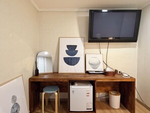 Habitación pequeña con escritorio de madera, refrigerador blanco compacto, televisor montado en la pared y una obra de arte abstracta enmarcada.