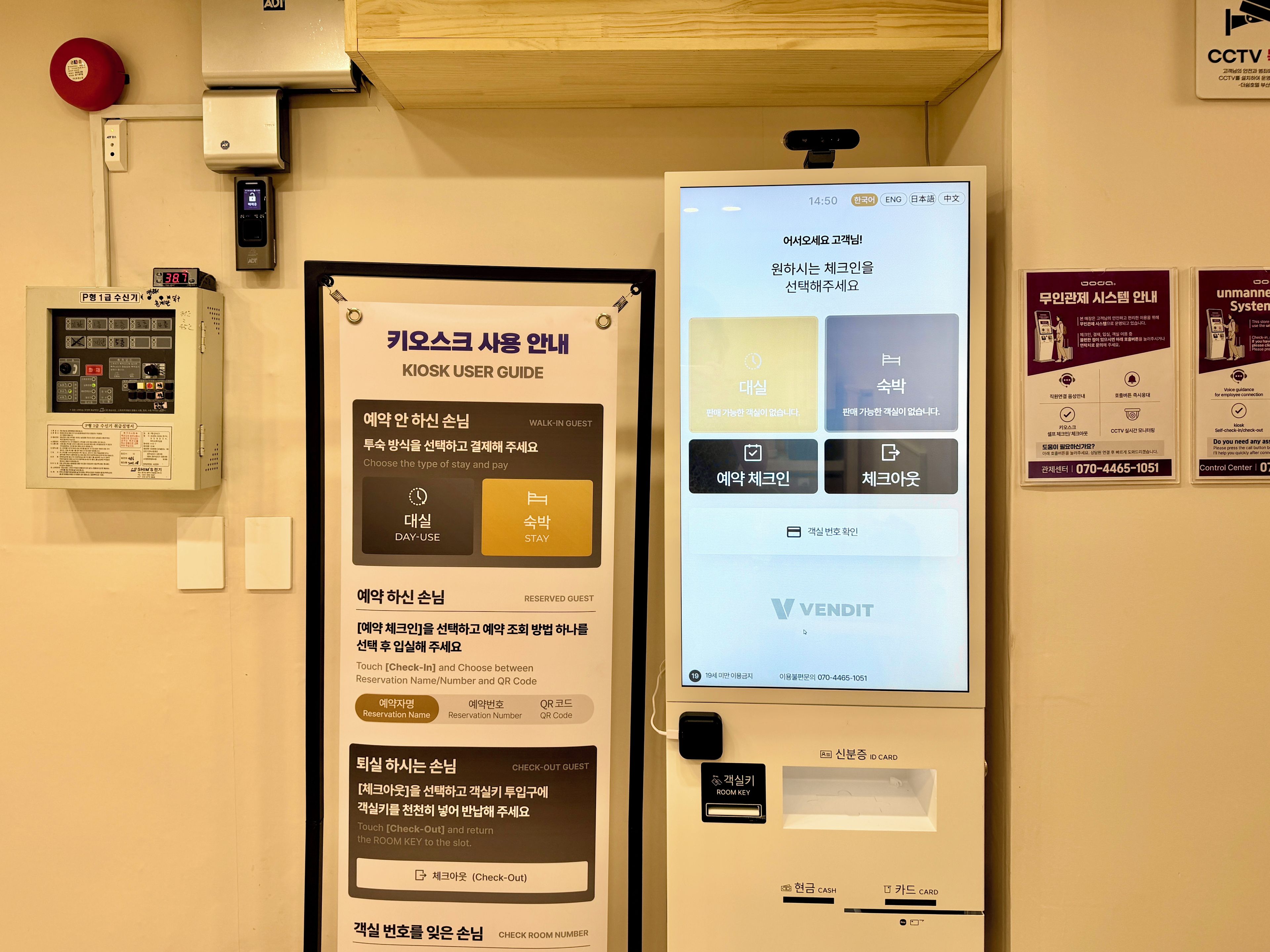 A hotel room with a digital kiosk for guest services, a control panel with various buttons, and a mounted fire alarm.