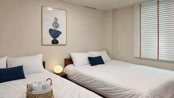 Habitación de hotel con dos camas, ropa de cama blanca, una cesta de mimbre con toallas, una obra de arte en la pared y una ventana con persianas.