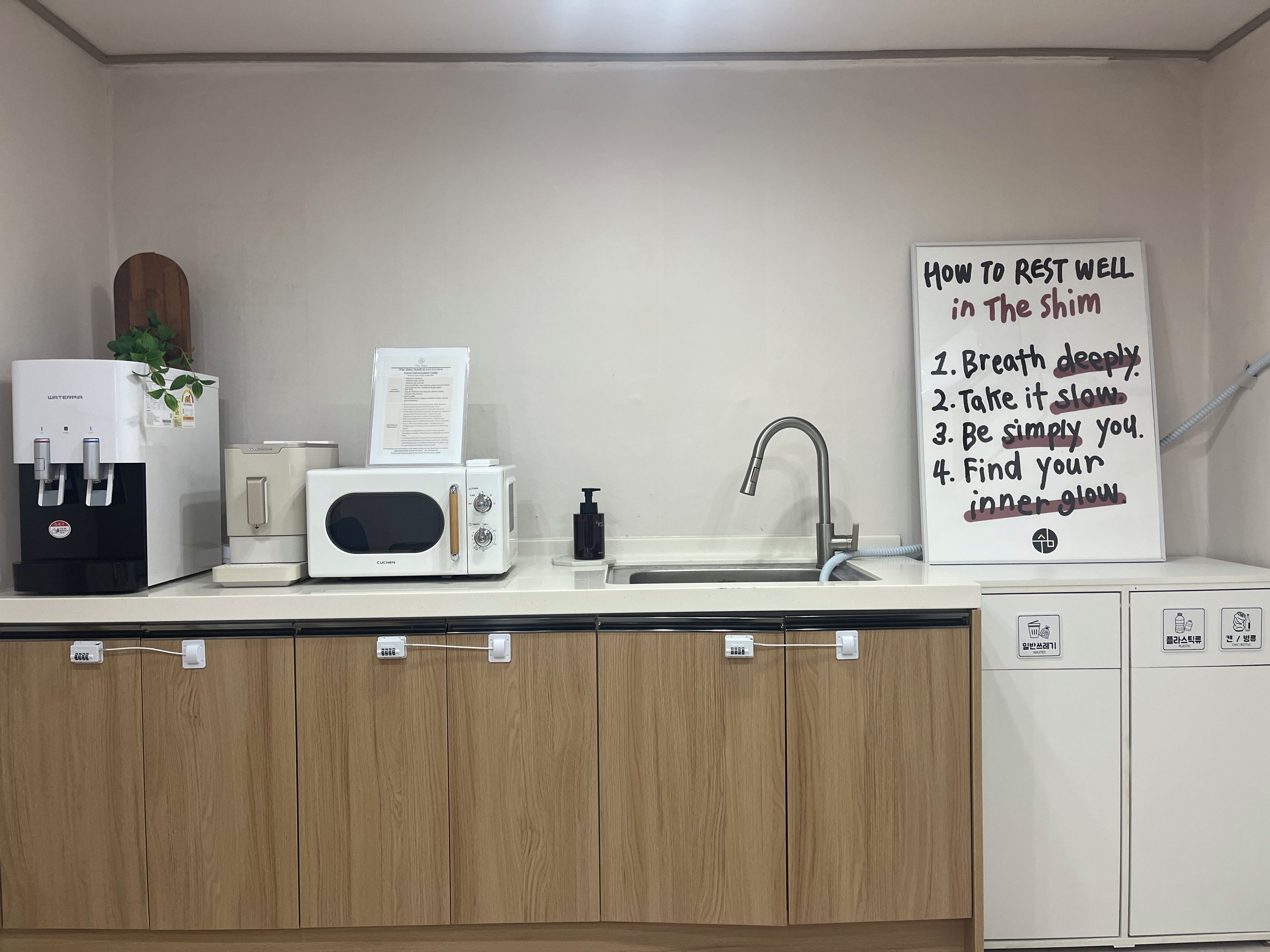 Una kitchenette equipada con dispensador de agua, microondas, cafetera y lavamanos. Hay un cartel con consejos para descansar bien.