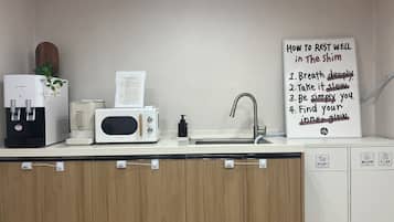 Una kitchenette equipada con dispensador de agua, microondas, cafetera y lavamanos. Hay un cartel con consejos para descansar bien.
