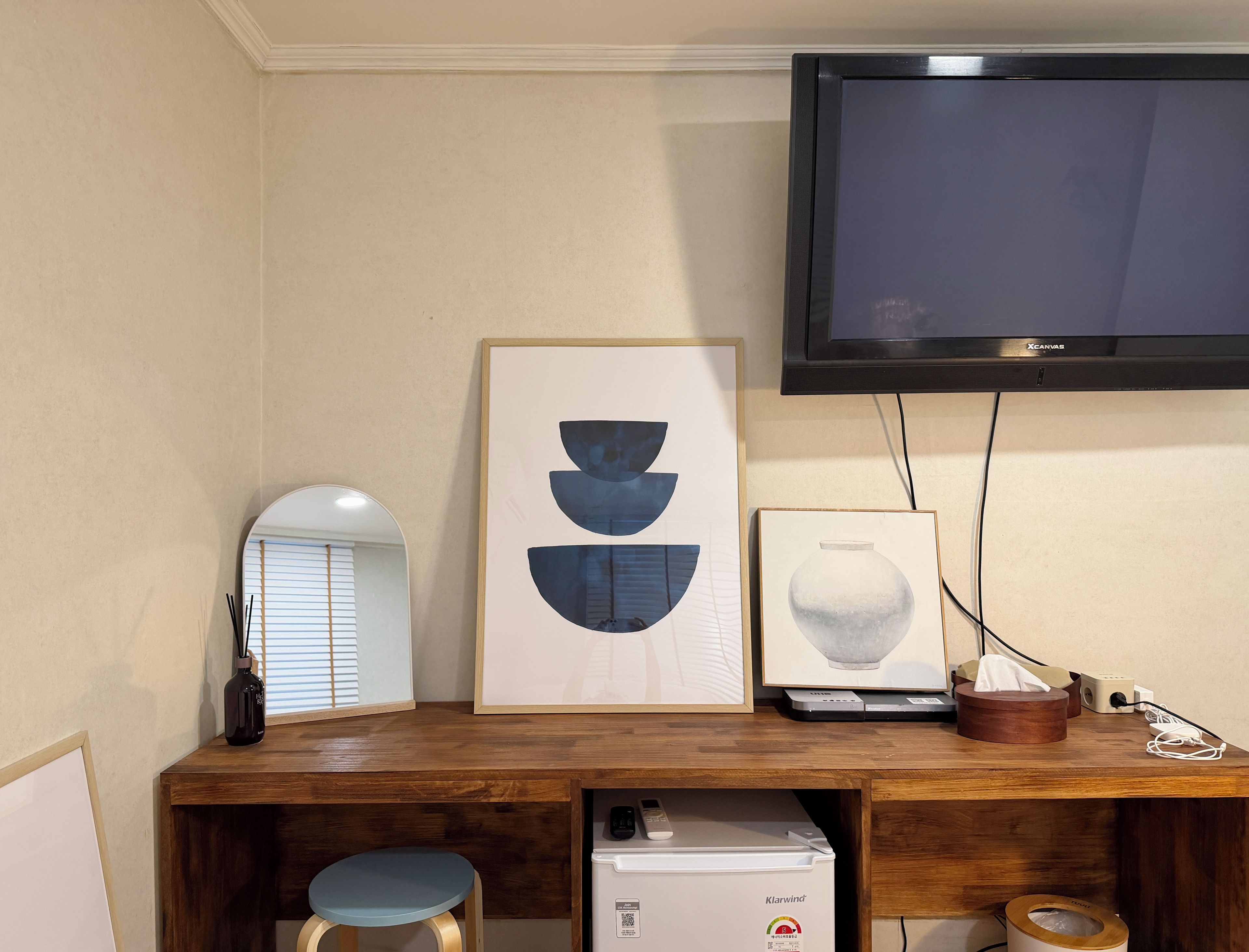 A room with a wooden desk, a flat-screen TV, a framed abstract painting, a vase, and a small refrigerator.