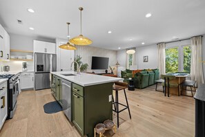 Een moderne keuken met een centraal eiland, een eetgedeelte met tafel en stoelen, een woonkamer met een bank, en een groot raam met gordijnen.