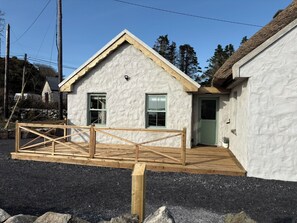 A small, single-story building with a sloped roof, a wooden deck, and a stone wall. - Gorse Cottage Connemara | Wild Atlantic Way | Sleeps 4 | Pet Friendly (Cashel)