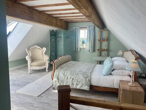 Ein Schlafzimmer mit schrägem Dach, einem Bett mit weißer Bettdecke, einer Holzleiter, einem Stuhl und einem kleinen Tisch mit Lampe.