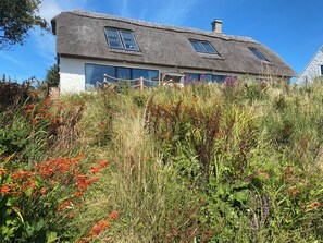 A house with a thatched roof, large windows, and surrounded by wildflowers. - Lios an Uisce Cottage Connemara | Wild Atlantic Way | Sleeps 6 | Pet Friendly (Cashel)