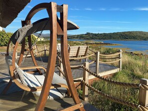 Ein Schaukelstuhl aus Holz mit Kissen, einem Seilabfangnetz und Blick auf einen See und Hügel.