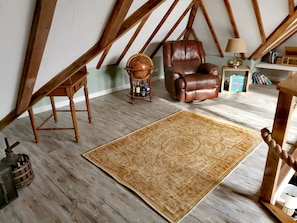 A loft-style room with exposed wooden beams, a leather armchair, a small wooden table with a lamp, and a patterned rug. - Lios an Uisce Cottage Connemara | Wild Atlantic Way | Sleeps 6 | Pet Friendly (Cashel)