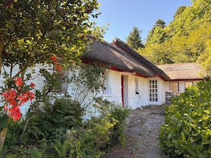 A traditional white building with a thatched roof, surrounded by lush greenery and a stone pathway leading to the entrance. - Mayfly Cottage in Oughterard | Wild Atlantic Way | Sleeps 6 | Pet Friendly (Cashel)