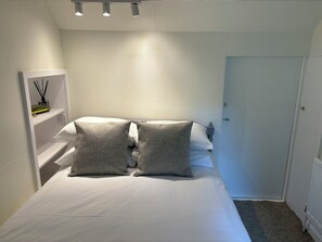 Une chambre à coucher avec un lit, deux oreillers, une étagère avec des livres et une bougie, et une porte.