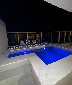 Ein Swimmingpool mit blau beleuchtetem Rand, umgeben von einer weißen Liegefläche und einer Sitzgelegenheit mit Blick auf den Himmel.