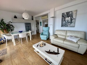 A modern living room with a white sofa, a coffee table with decorative items, a dining area with a white table and chairs, and a large painting on the wall. - Luxury Penthouse with Panoramic Sea Views (Vilanova i la Geltru)