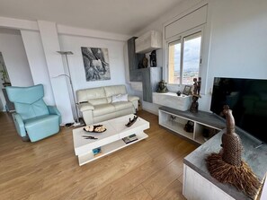 A modern living room with a sofa, a blue armchair, a coffee table, and a TV. - Luxury Penthouse with Panoramic Sea Views (Vilanova i la Geltru)