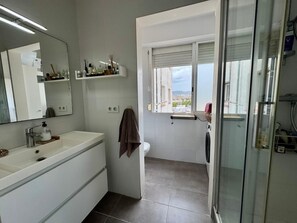 Ein Badezimmer mit weißem Waschtisch, einem Spiegel, einem Handtuchhalter, einem Fenster mit Aussicht und einer Duschkabine aus Glas.