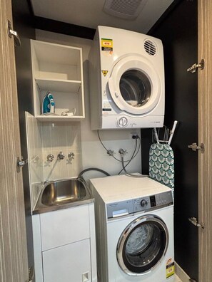 A compact laundry room with a front-loading washing machine, a small sink, and a shelf with cleaning supplies. - Zetland 3BR·Sleeps 7 | Parking · 4min to Central (Zetland)