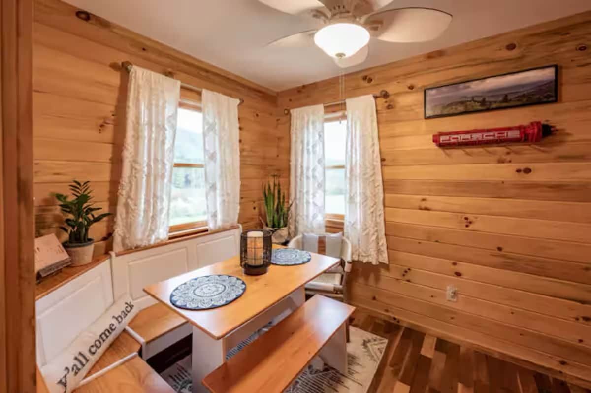 En inredning med träpanel, ett matbord, två fönster med gardiner, en takfläkt och en inramad tavla på väggen.