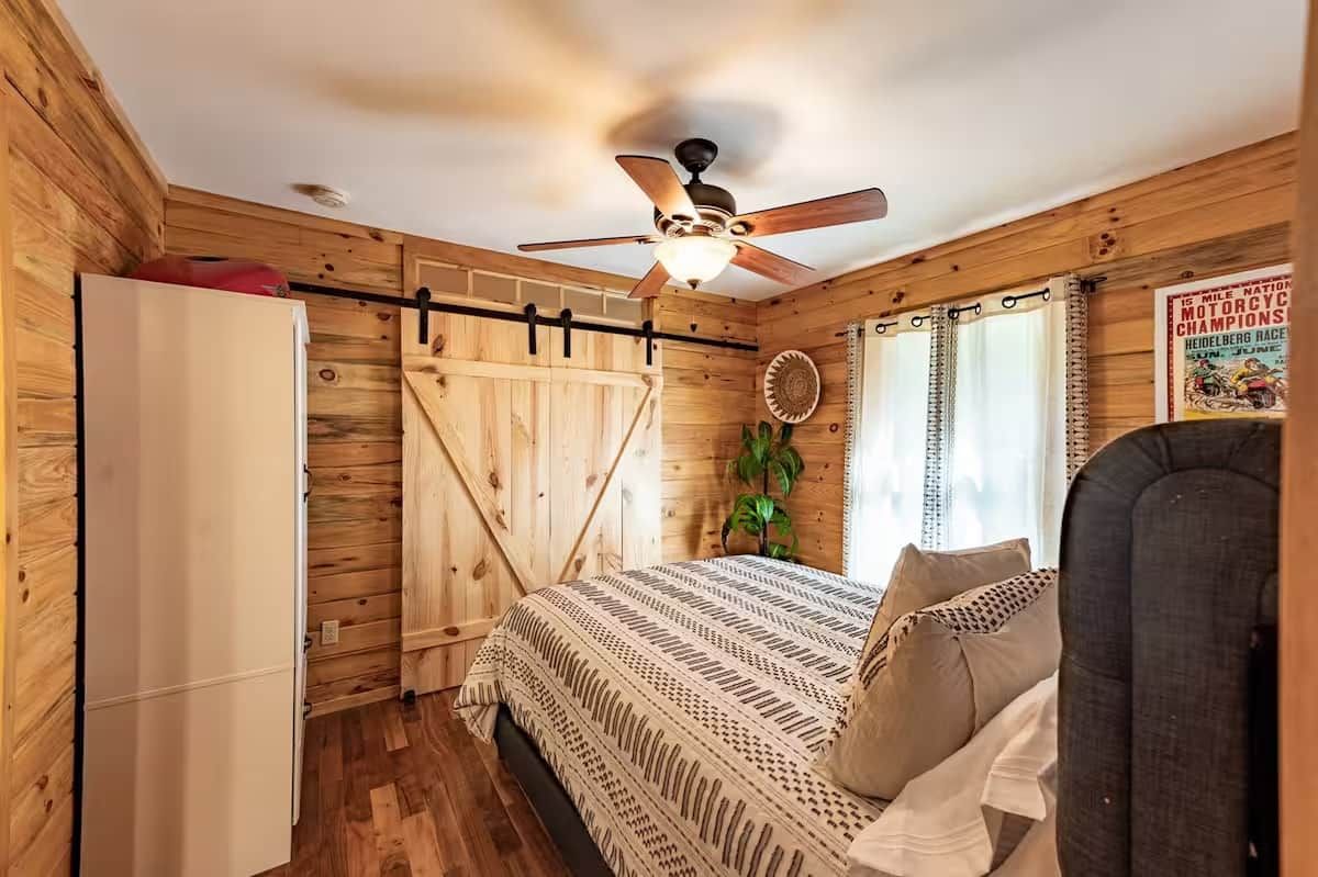 Un dormitorio tipo cabaña de madera con una cama, un ventilador de techo, una ventana con cortinas y un cartel en la pared.