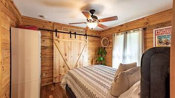 Un dormitorio tipo cabaña de madera con una cama, un ventilador de techo, una ventana con cortinas y un cartel en la pared.