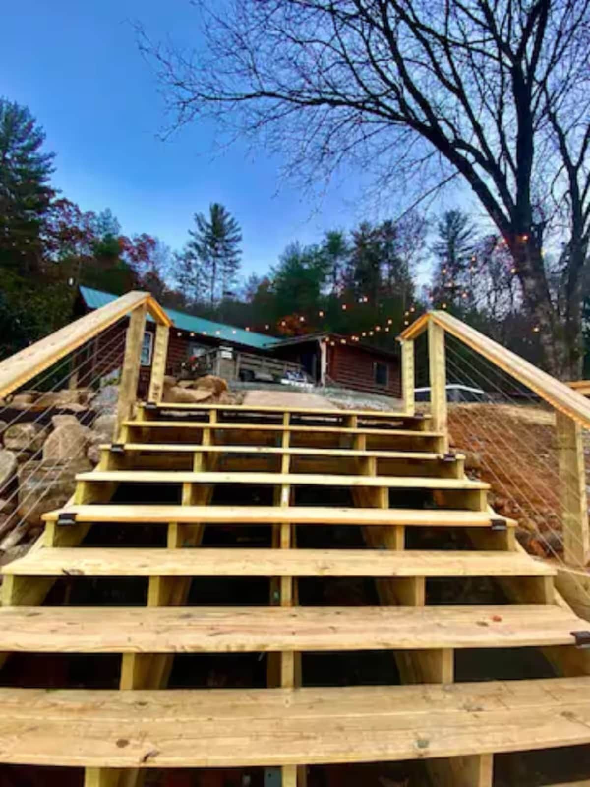 A wooden staircase leading to a cabin with a metal railing, surrounded by trees and a clear sky.