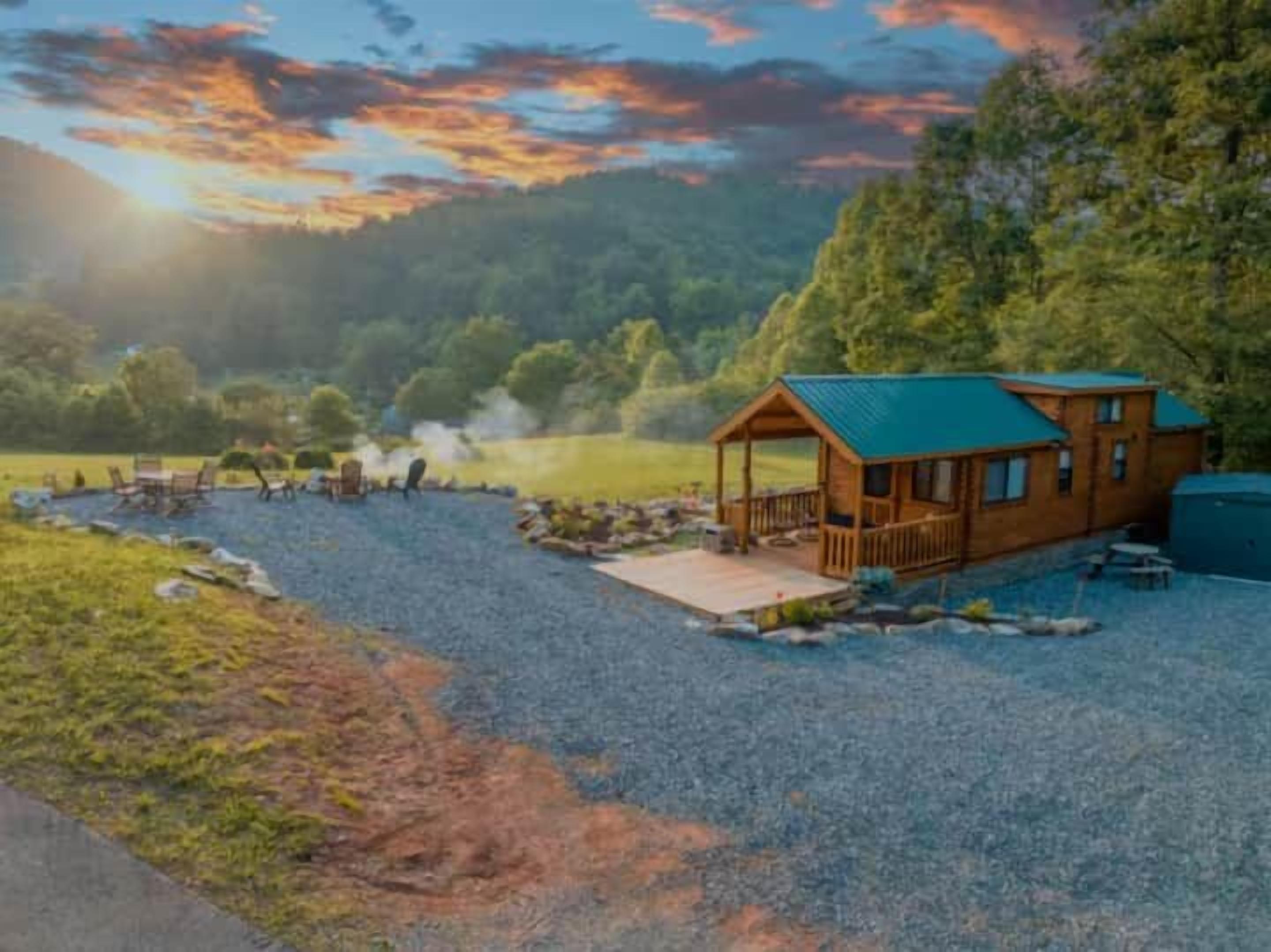 A cabin with a deck, surrounded by a gravel area and a body of water, with trees and a sunset in the background.