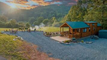 A cabin with a deck, surrounded by a gravel area and a body of water, with trees and a sunset in the background.