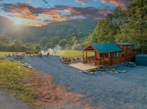 A cabin with a deck, surrounded by a gravel area and a body of water, with trees and a sunset in the background.