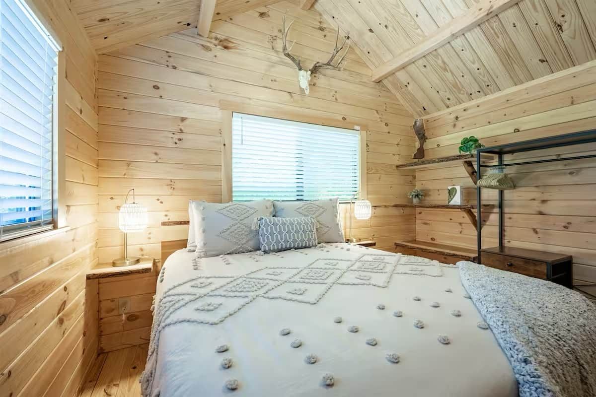 Ett sovrum i en stuga med träpanel, inrett med en säng, två sängbord, ett fönster och ett hjorthuvud som hänger i taket.
