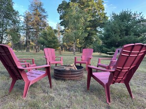 Eine Gruppe Adirondack-Stühle, die um eine Feuerstelle in einer grünen Außenanlage mit Bäumen im Hintergrund angeordnet sind.