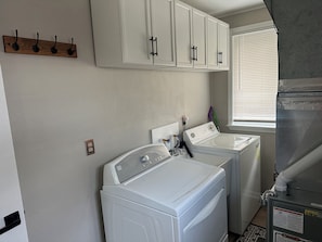 A laundry room with two white front-loading washers and dryers, a wooden coat rack, and a window with blinds. - Cozy Cabin/ORV/Fishing/Fire Pit/Dog Friendly (Irons)