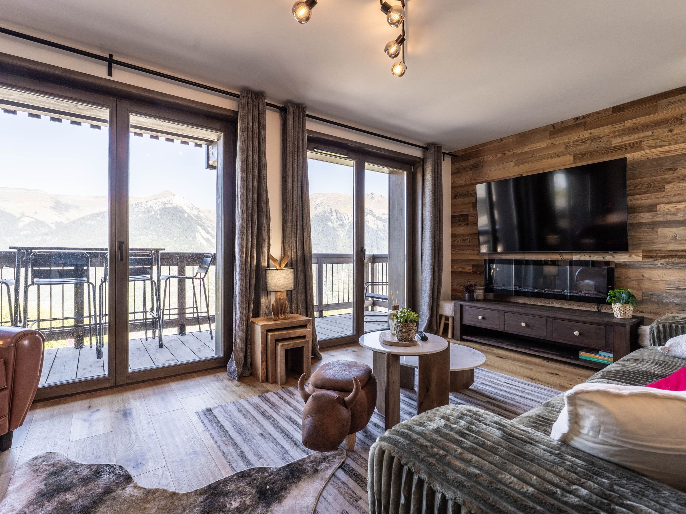 Une chambre d’hôtel moderne dotée d’une grande télévision, de murs en bois, d’un balcon avec des chaises et d’une vue sur les montagnes.