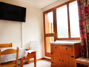 Ein Zimmer mit einem Holztisch, einem Stuhl, einem an der Wand montierten Fernseher, einem Fenster mit Vorhängen und einem Holzschrank.