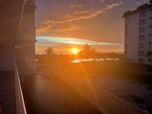 En solnedgång sedd från en balkong med utsikt över ett bostadsområde, mot en klar himmel och en sol som står lågt.