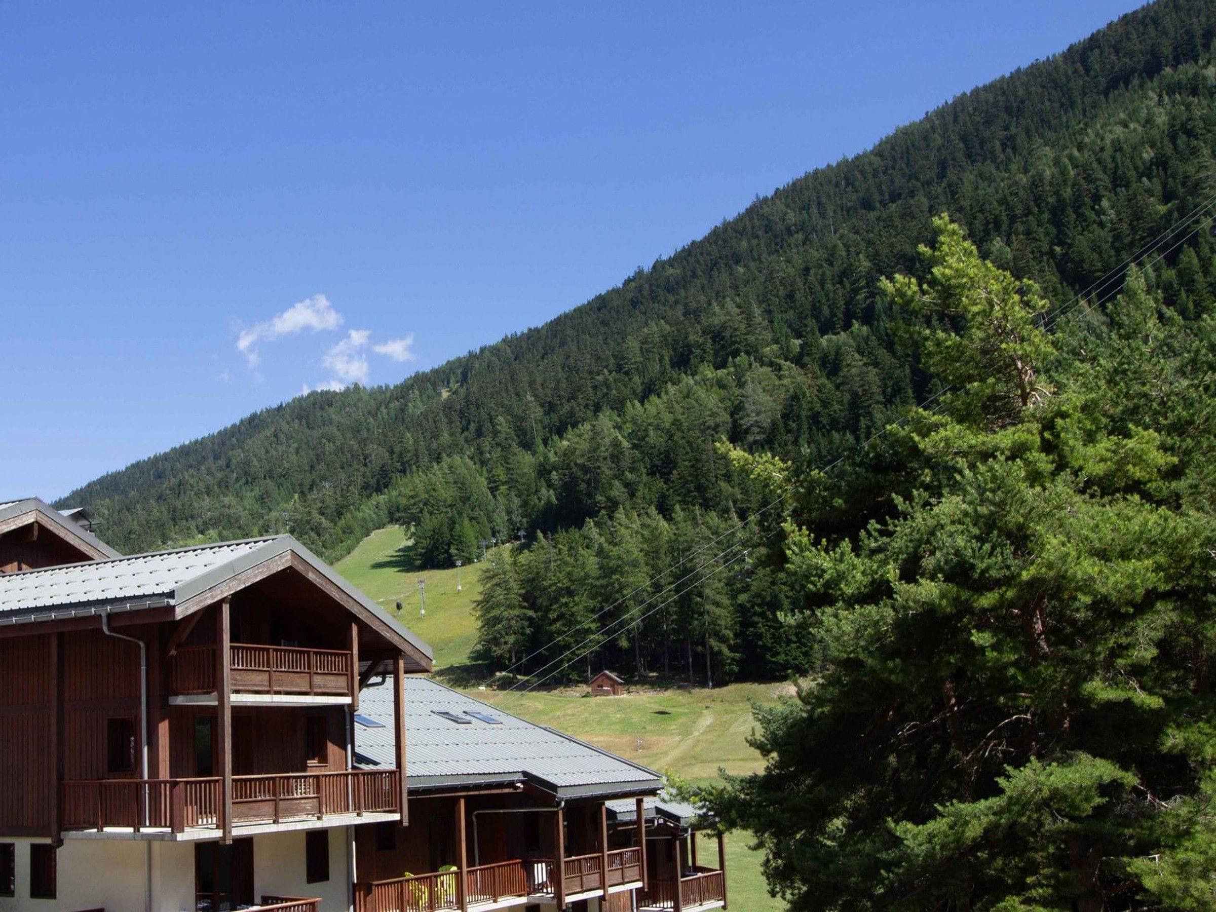 A two-story wooden building with balconies, set against a backdrop of a forested mountain.