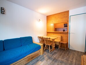Ein kompakter Wohnraum mit blauem Sofa, Esstisch aus Holz und Stühlen, einer Küchenzeile mit Holdekor-Schränken und einer weißen Tür.