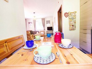 Ein Holztisch mit einem blauen und grünen Geschirrset, einem roten Wasserkocher und einer weißen Tasse. Eine hölzerne Bank befindet sich links, und ein Kühlschrank rechts.