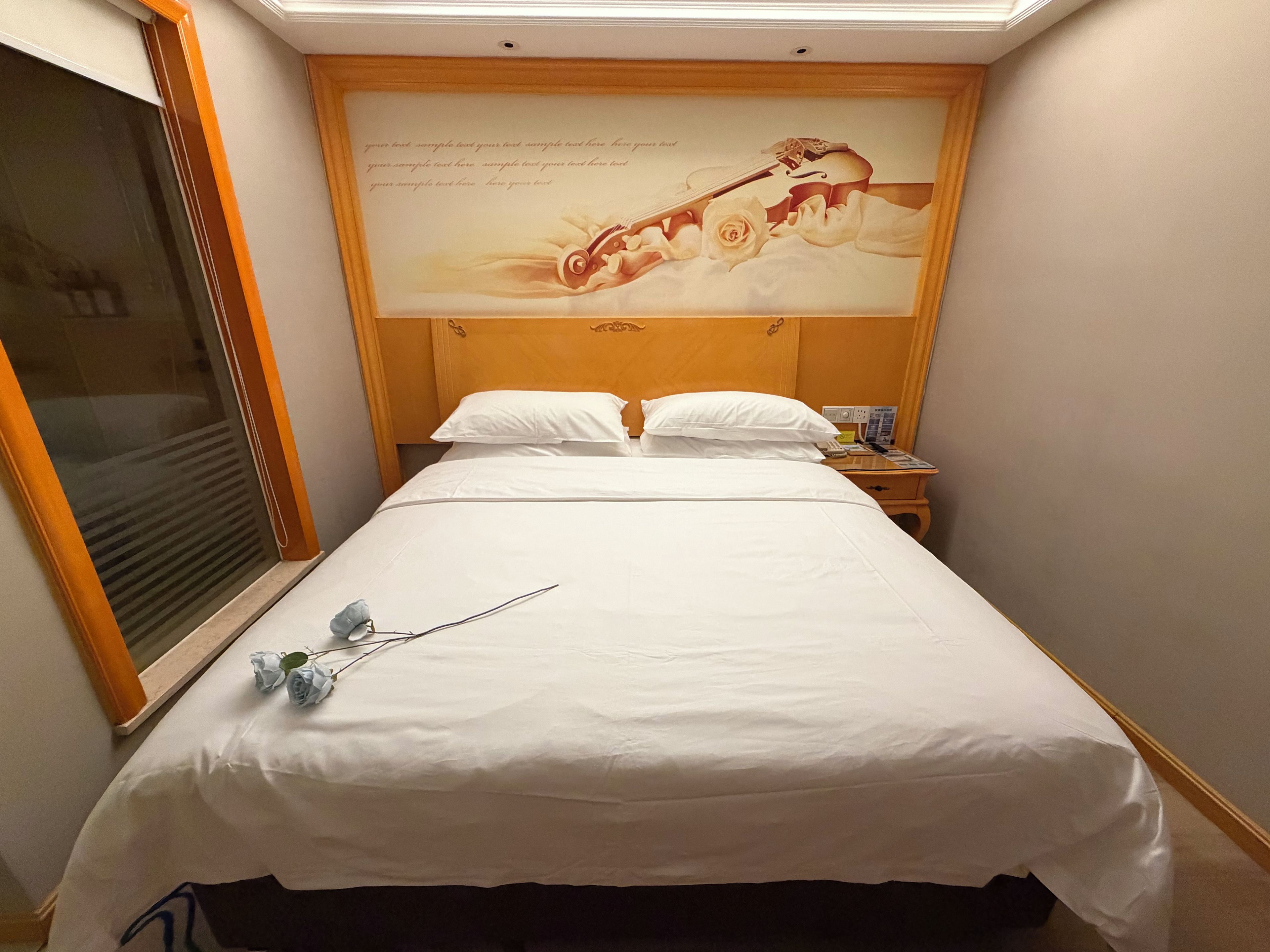 A hotel room with a large bed, a nightstand, a wall-mounted artwork, and a window with blinds.