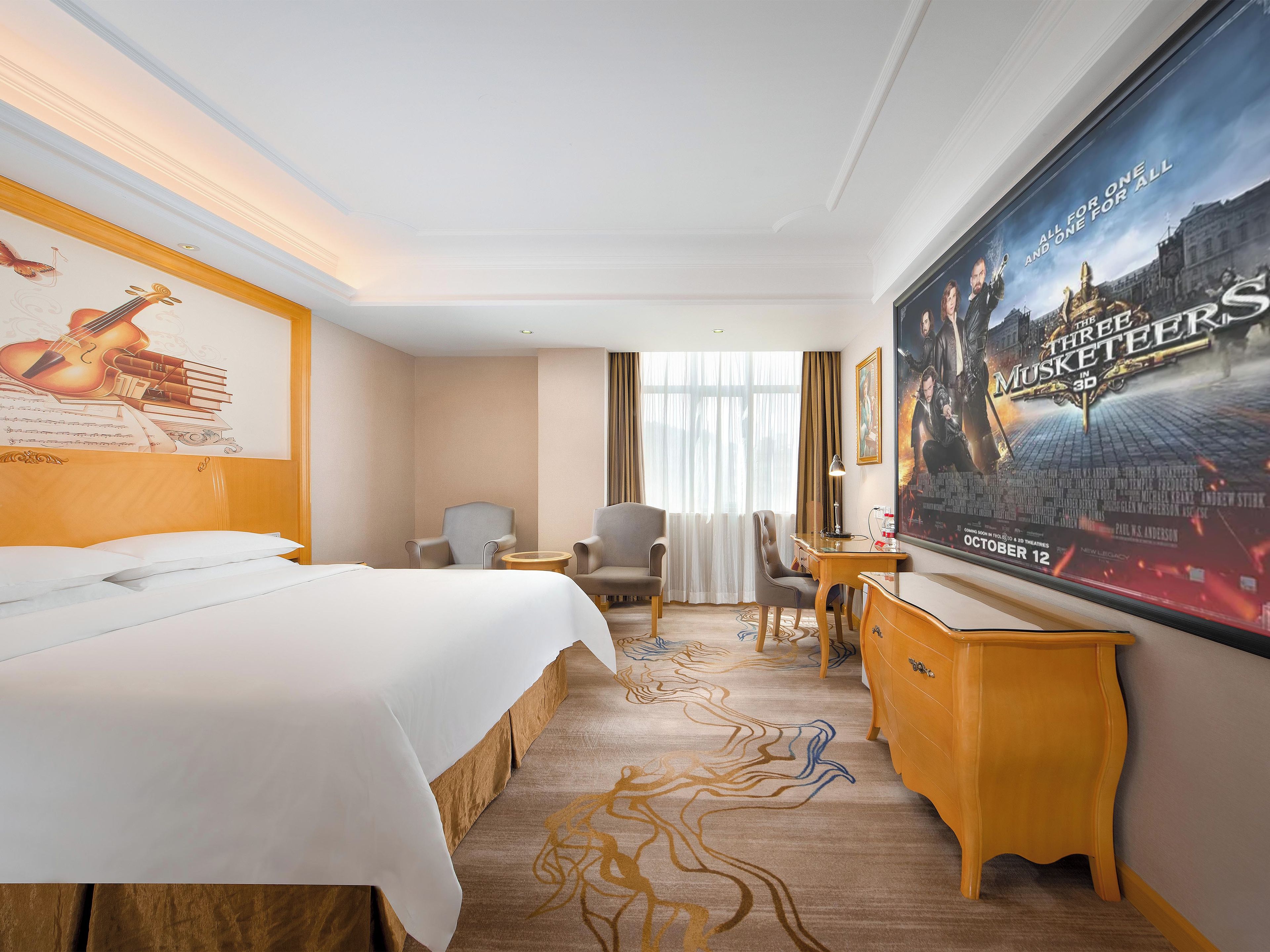 Une chambre d’hôtel avec un lit, deux fauteuils, un bureau, une grande affiche et une fenêtre avec des rideaux.