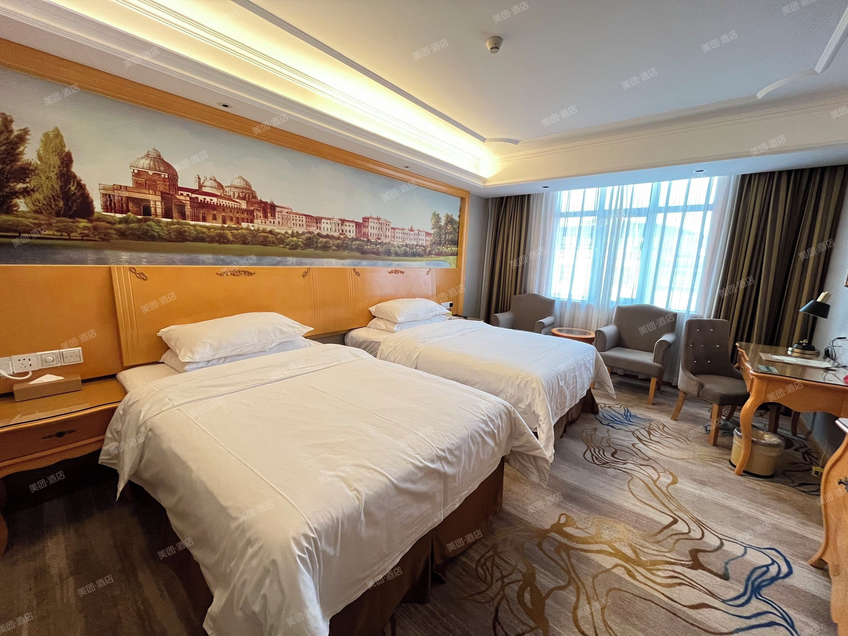 Habitación de hotel con dos camas, un escritorio, una silla y un amplio ventanal con cortinas.
