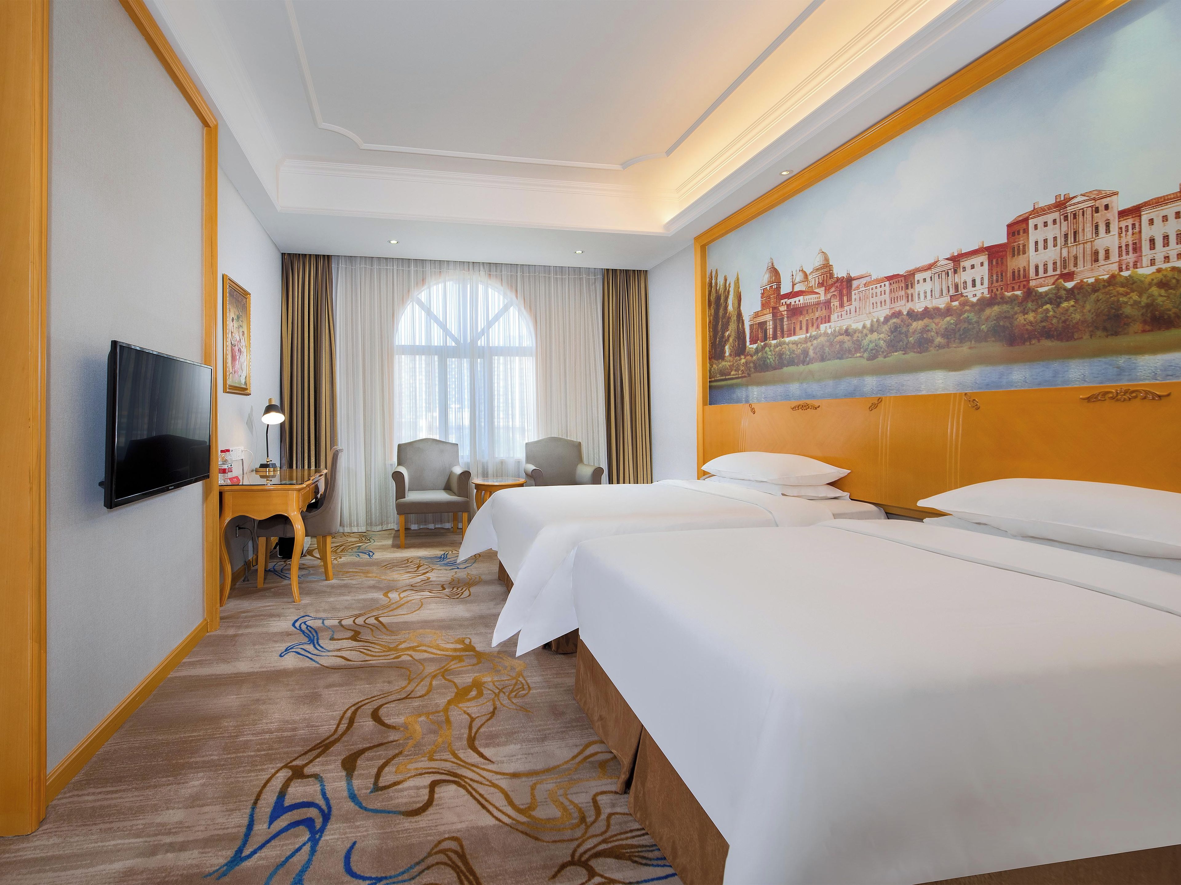 Une chambre d’hôtel avec deux lits, un bureau avec une chaise, une télévision et une grande fenêtre avec des rideaux.